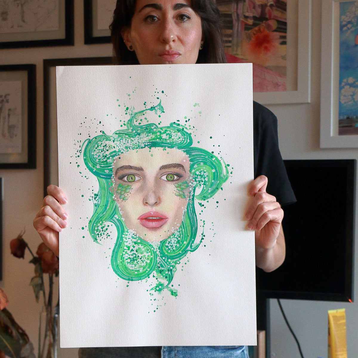 Artist Paula Di Marco holding her original watercolor painting 'Mermaid'—a surreal portrait of a woman with luminous green eyes and flowing sea-green hair, symbolizing mystical femininity and emotional healing. Photographed in her studio, this hand-drawn piece is part of her expressive watercolor wall art collection.