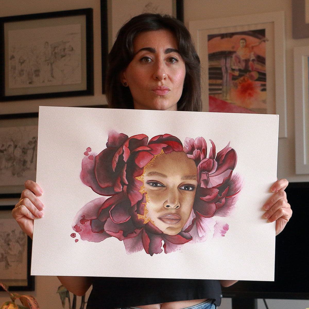 Artist Paula Di Marco holding her original watercolor painting 'Pensive Bloom', a surreal portrait of a woman framed by deep red petals and golden accents. Captured in her studio, this emotionally charged artwork is part of Paula’s mystical watercolor collection focused on reflection and healing.
