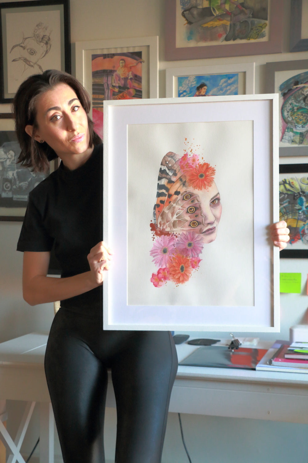 Paula Di Marco holding a framed surrealist watercolor portrait featuring a woman's face merged with vibrant flowers and a butterfly wing — a hand-drawn giclée print symbolizing emotional expression.