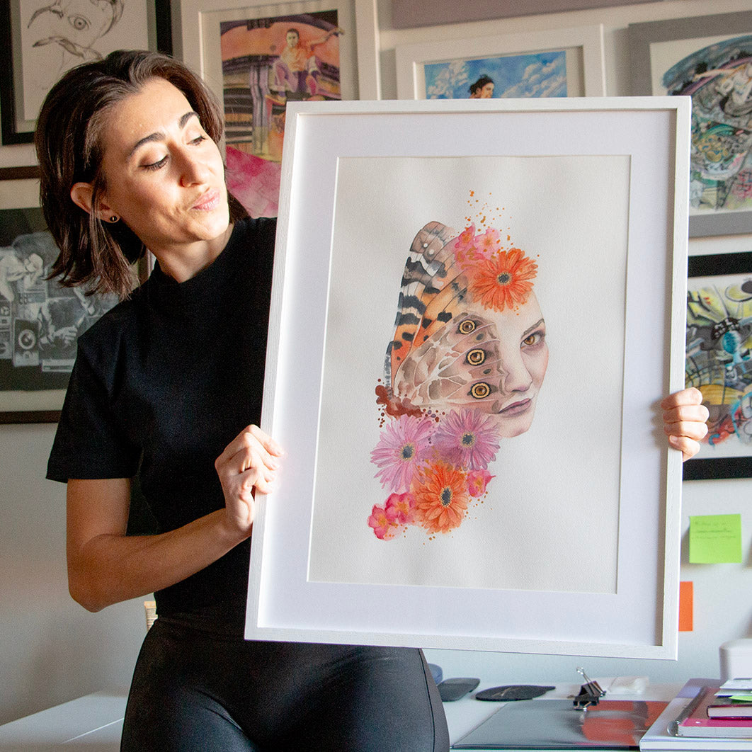 Artist Paula Di Marco holding her original watercolor painting 'Look Closer'—a surreal, emotionally charged portrait blending a woman’s face with butterfly wings and vibrant florals. Photographed in her studio, this framed piece is part of her mystical collection of watercolor wall art for emotional healing and introspection.