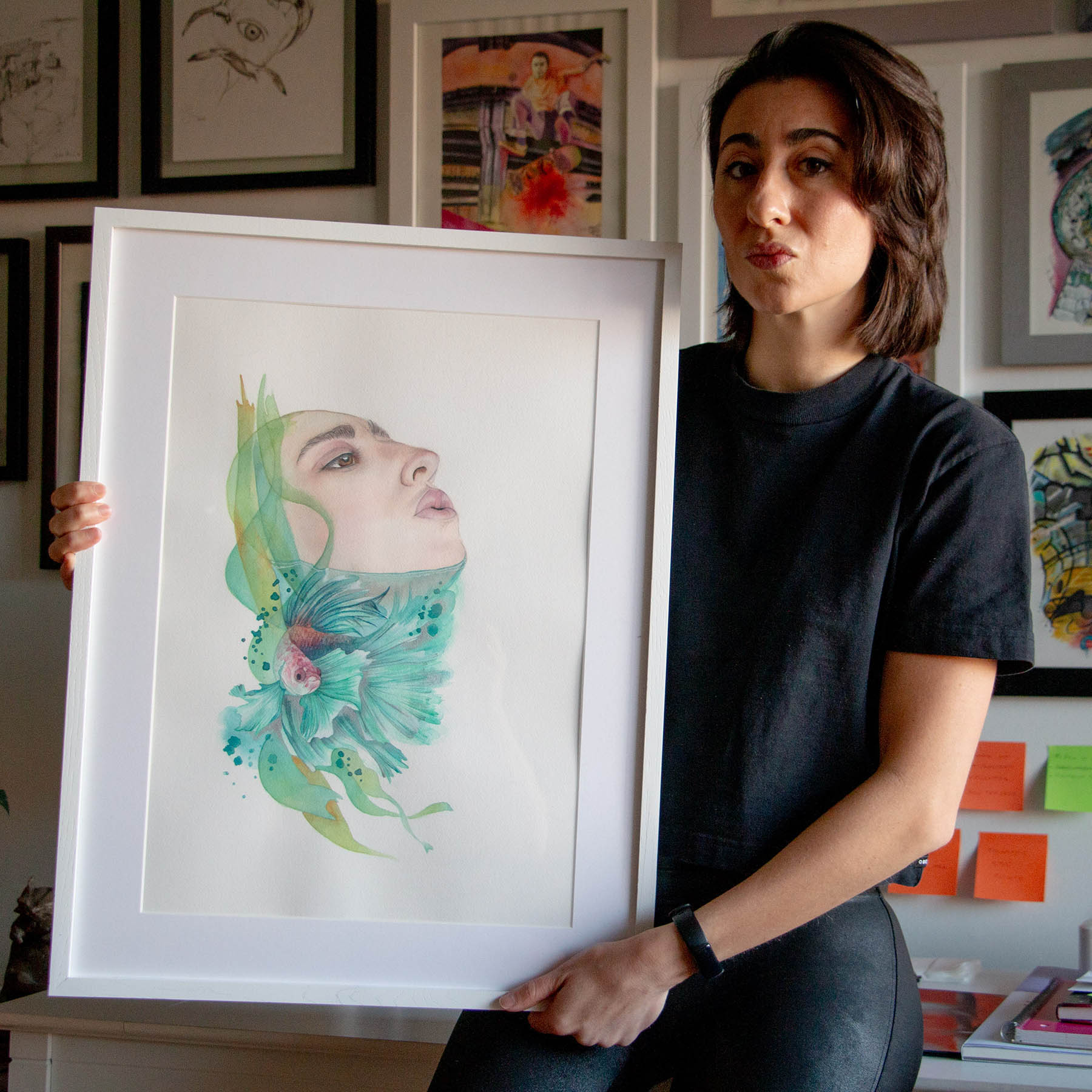 Artist Paula Di Marco holding her original framed watercolor painting 'A Breath of Air'- a surreal, emotionally charged portrait blending human and aquatic elements. Photographed in her studio, this piece is part of her mystical art collection supporting emotional healing and introspection.