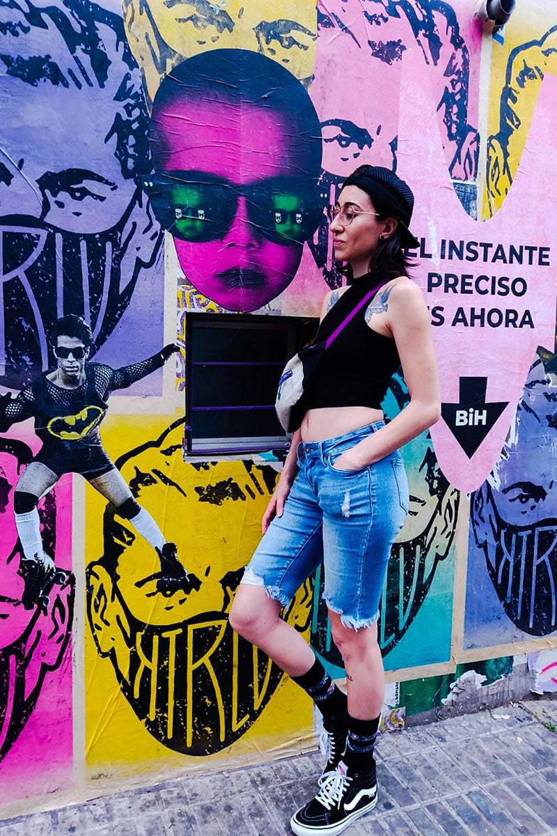 Artist Paula Di Marco standing against a vibrant surrealist mural wall in casual streetwear — blending urban expression with her identity as a creator of emotionally charged watercolor art.