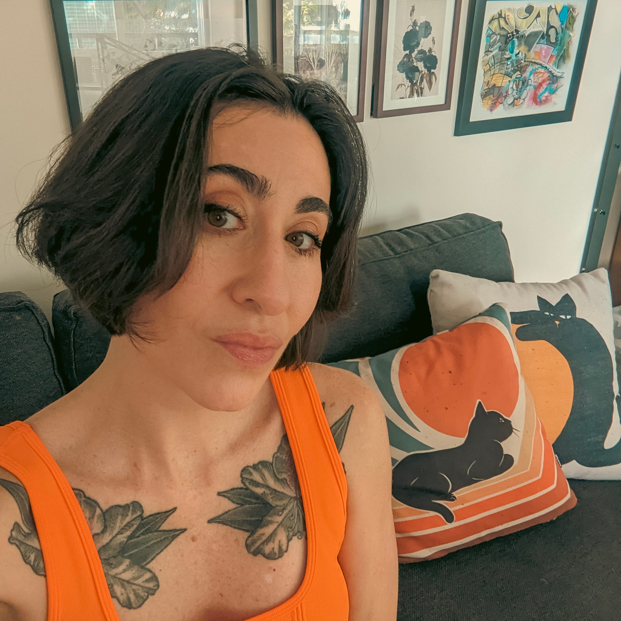 Artist Paula Di Marco with botanical tattoos sitting in her art studio , framed artwork, and cat-themed pillows in the background