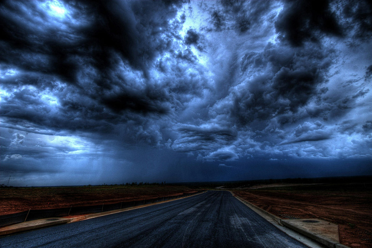A dark stormy sky with swirling blue-black clouds looms over a long, empty road through barren land.