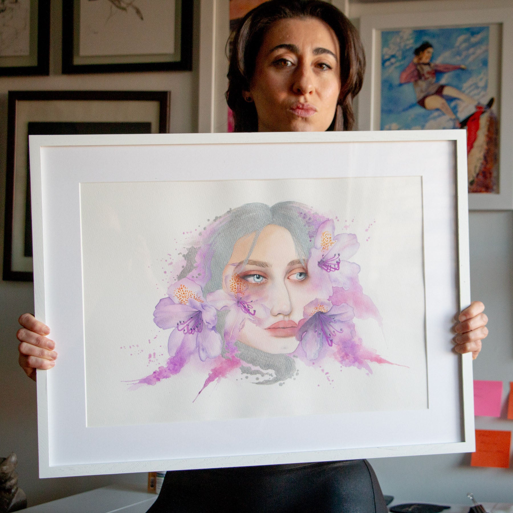 Artist Paula Di Marco holding her original watercolor painting 'Let’s Sweep'—a surreal portrait of a woman surrounded by soft purple flowers and brushwork, evoking grief, vulnerability, and emotional healing. Part of Paula’s mystical collection of emotionally charged watercolor wall art.