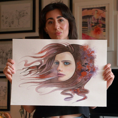 Artist Paula Di Marco holding her original watercolor painting 'Release'—a surreal portrait of a woman with flowing hair blending into warm, abstract brushstrokes. Captured in her studio, this emotionally charged artwork is part of her mystical watercolor wall art collection focused on grief, transformation, and healing.